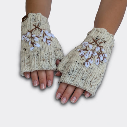 Mitaines écrues tricotées main, portées à plat. Le fond moucheté présente de petits fils noirs à effet tweed. Des fleurs de coton blanches et des tiges brunes décorent le dessus des mains.