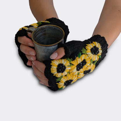 Mitaines noires tenant une tasse, décorées de fleurs de tournesol jaunes et blanches avec perles noires au centre.