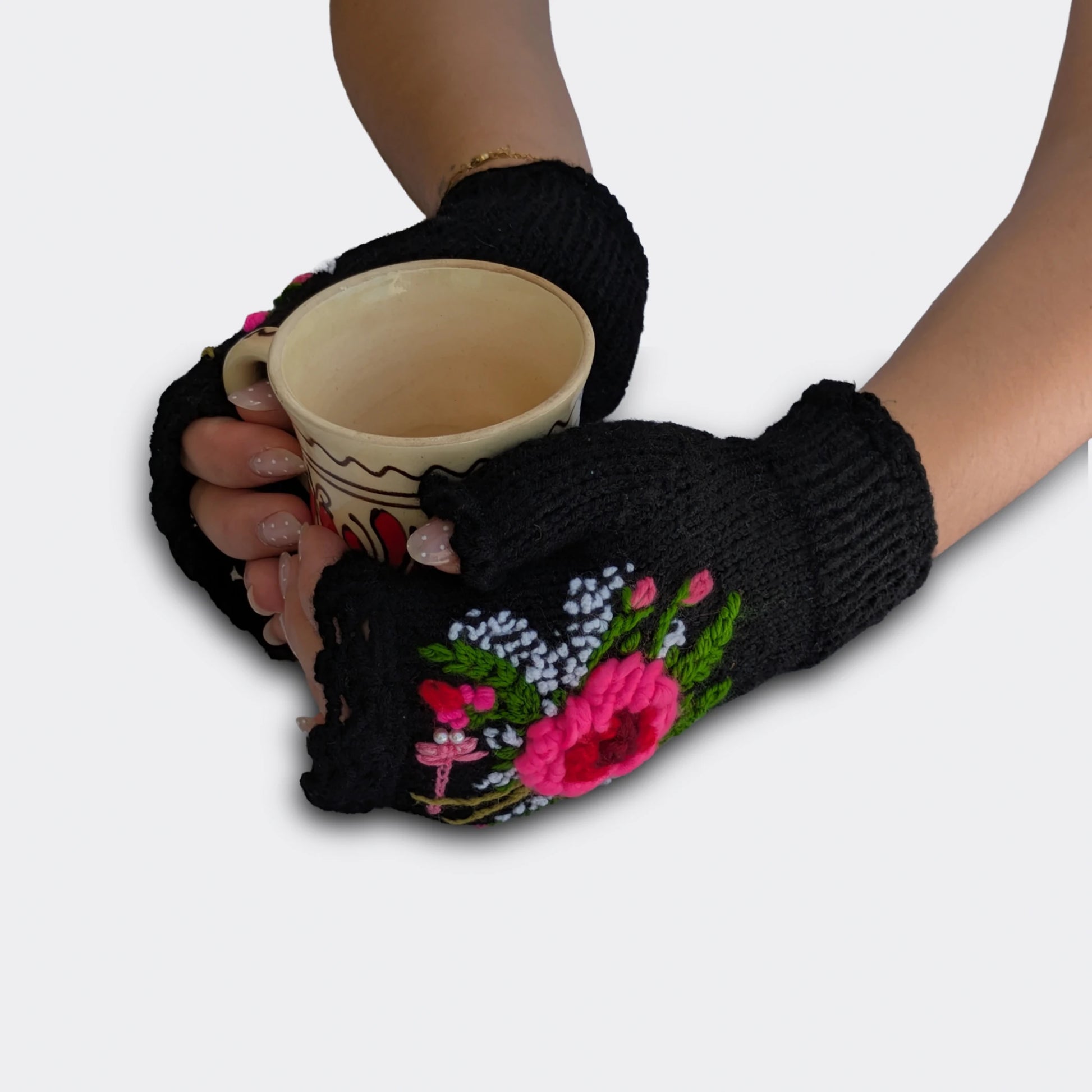Mitaines femme noires portées avec une tasse entre les mains. Le décor associe fleur rose, libellule brodée et petits détails blancs sur une base tricotée main. Le bord supérieur est festonné.