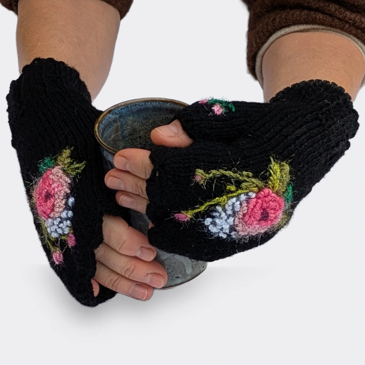 Des mains tenant une tasse portent des mitaines noires ornées de roses crochetées aux reflets scintillants. Des tiges vert olive, de petits boutons roses et des touches blanches complètent le décor.