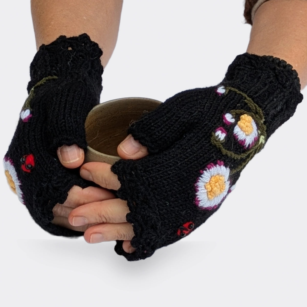 Des mains tenant une tasse portent des mitaines noires à fleurs blanches et jaunes. Une petite coccinelle rouge accompagne les pâquerettes et leurs boutons floraux sur le dos des mains.