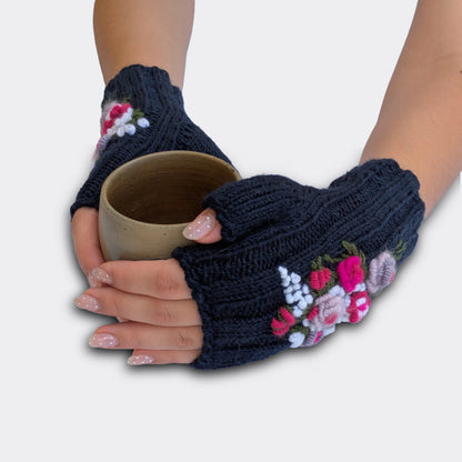 Mitaines femme bleu nuit portées avec une tasse entre les mains. Le décor présente des roses anciennes en relief, des petites fleurs blanches et un feuillage brodé. La paire est tricotée main dans une maille côtelée souple.