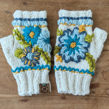 Mitaines blanc cassé tricotées main, décorées de fleurs bleues et turquoise reliées par des broderies vertes. Posées à plat.