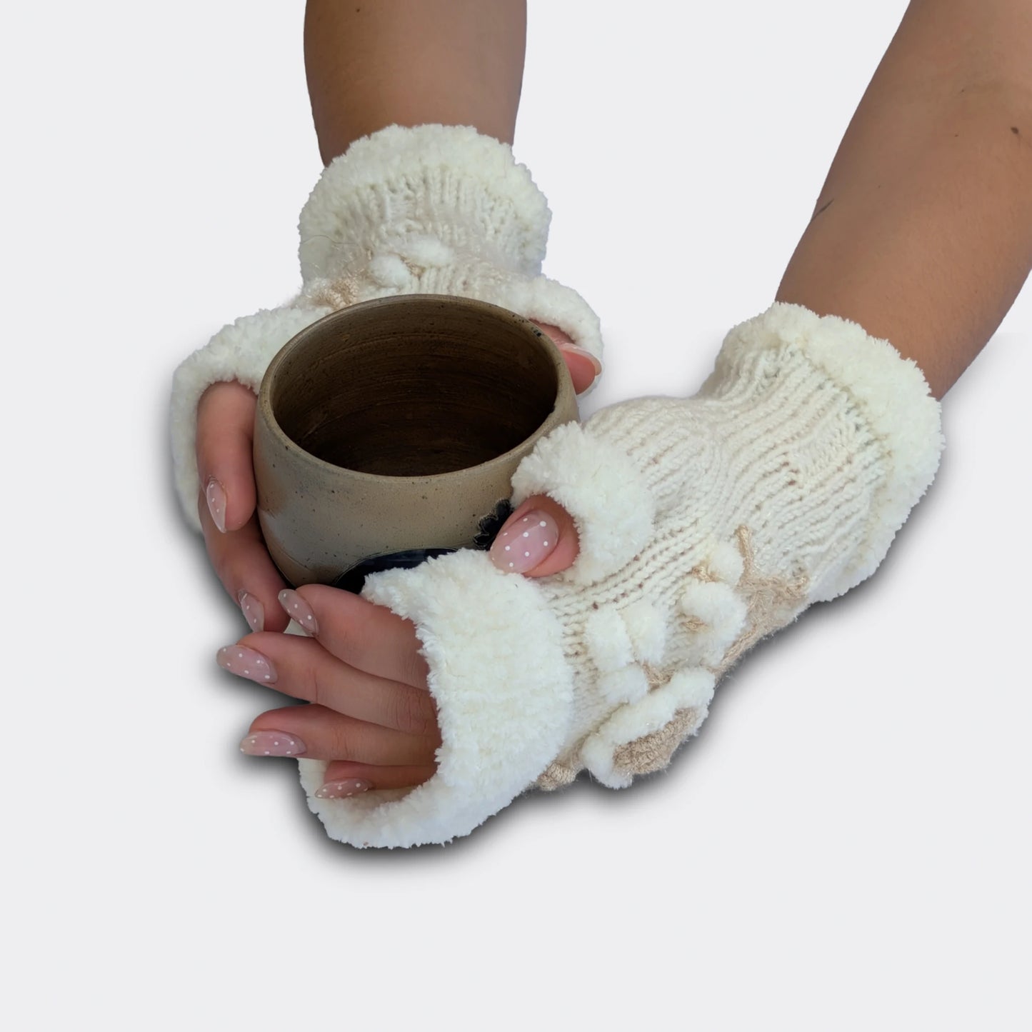 Mitaines florales blanches tenant une tasse. La maille tricotée main reçoit une fleur en relief, des petites boules blanches et une tige écrue brillante. Les bords pelucheux donnent une finition douce au modèle.