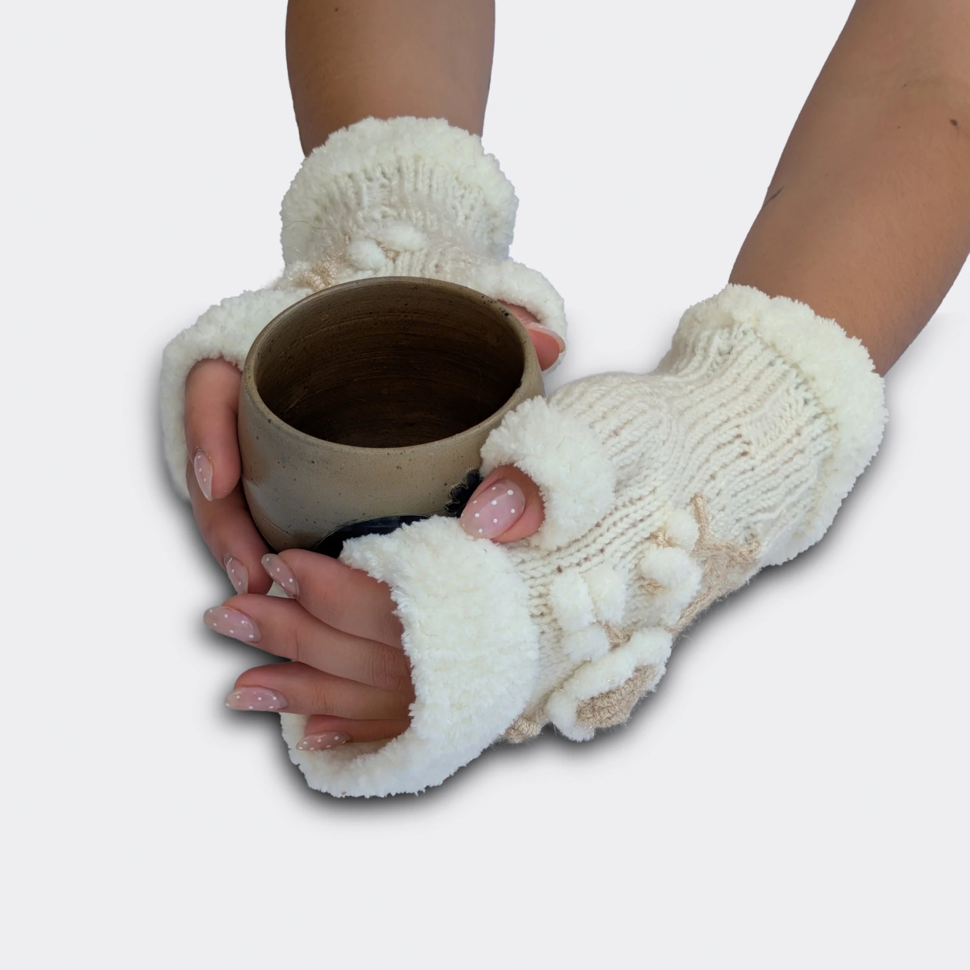 Mitaines florales blanches tenant une tasse. La maille tricotée main reçoit une fleur en relief, des petites boules blanches et une tige écrue brillante. Les bords pelucheux donnent une finition douce au modèle.