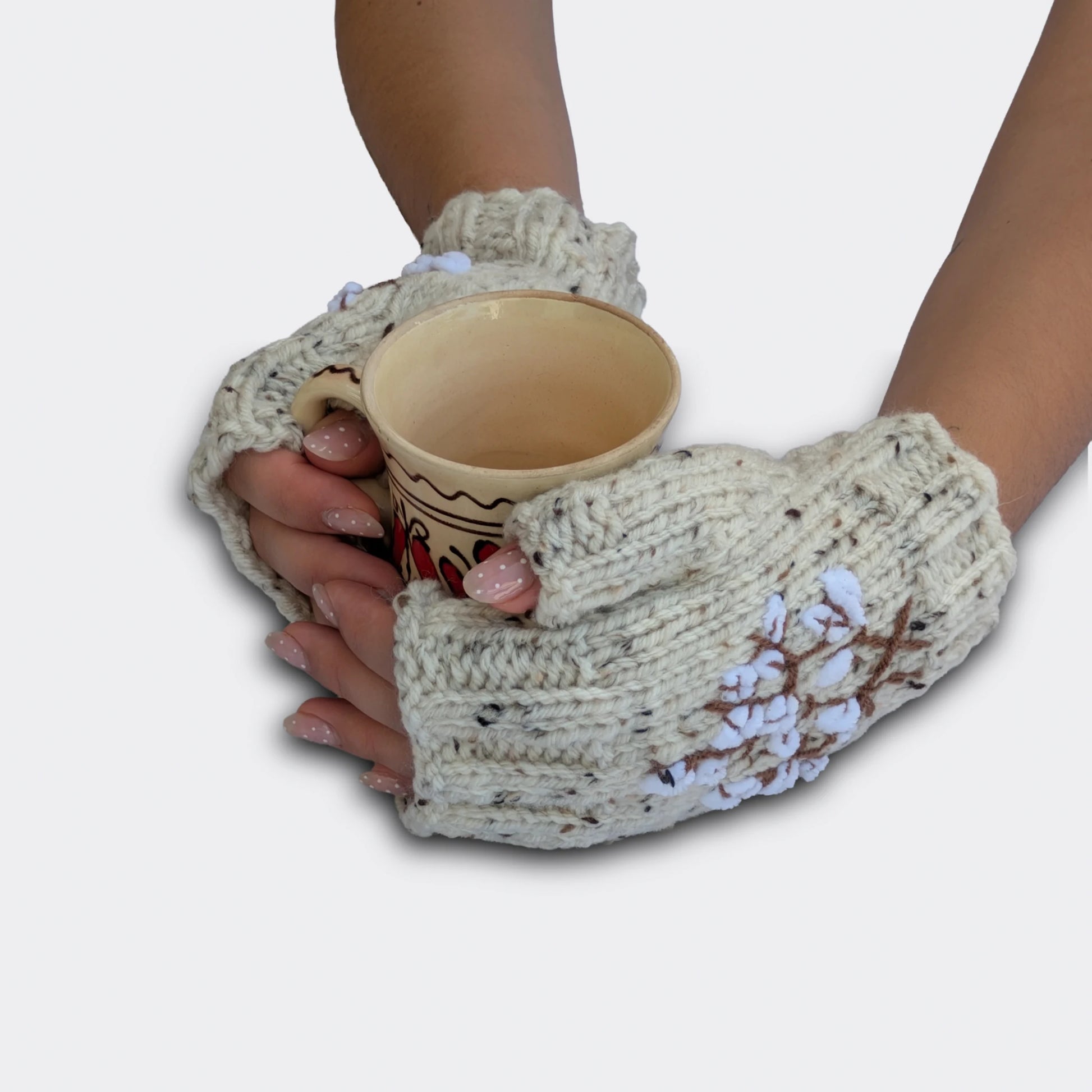 Mitaines écrues tenant une tasse, avec branches de coton blanches sur fond moucheté. Le tricot présente de petits fils noirs à effet tweed. Les tiges brunes brodées soutiennent les fleurs délicates.