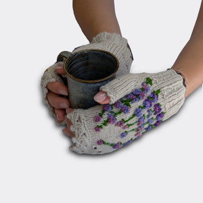 Mitaines femme gris clair portées avec une tasse entre les mains. Le décor mêle fleurs butinées, nuances de mauve et finition festonnée sur le bord supérieur. Travail artisanal.
