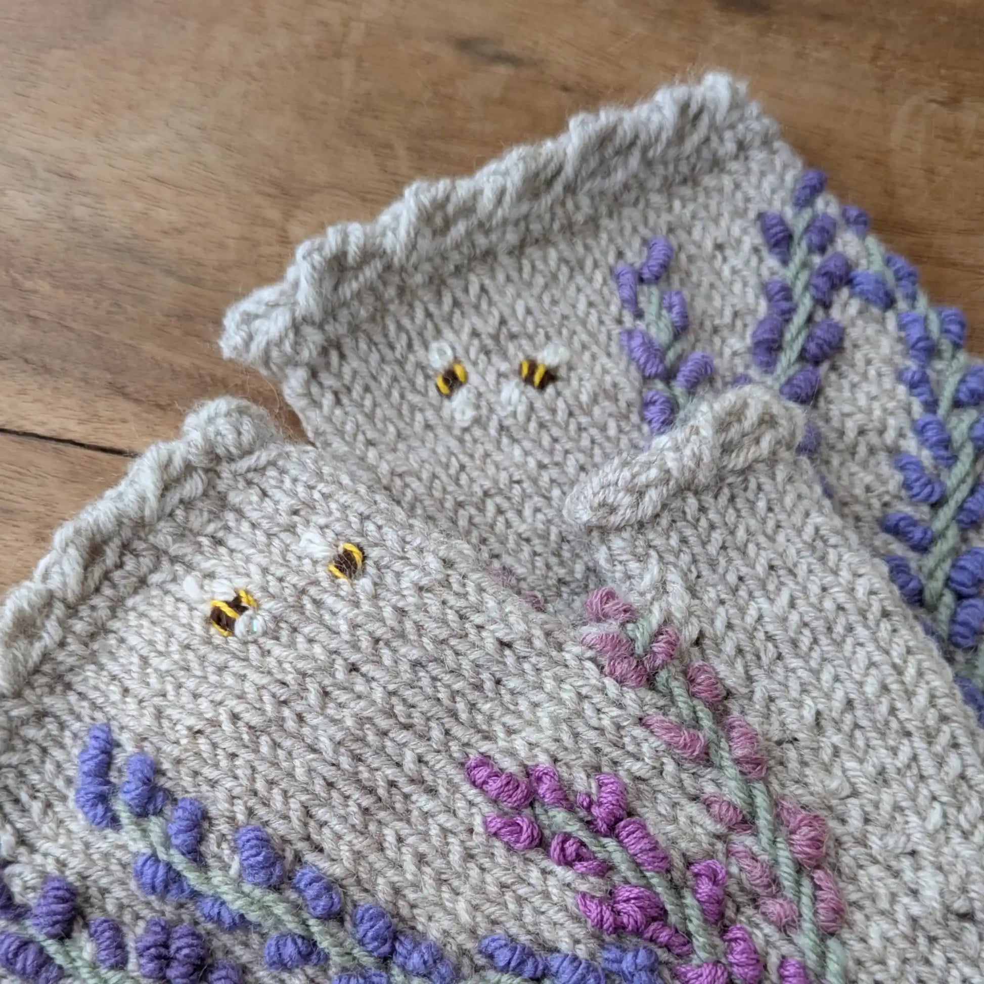 Gros plan sur des mitaines gris perle tricotées main, avec lavandes mauves et petites abeilles brodées.