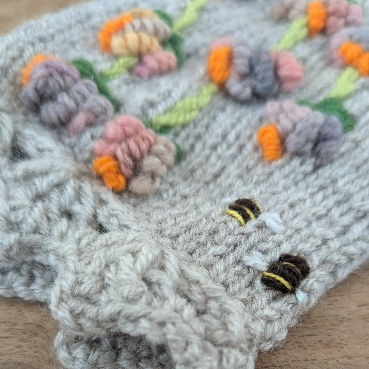 Composition florale multicolore sur des mitaines artisanales en tricot gris clair. Les fleurs en relief aux touches orange vif sont accompagnées de petites abeilles rayées, près de la bordure festonnée.