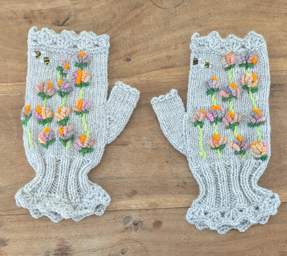 Mitaines femme grises tricotées à la main, présentées à plat sur une table en bois. Des fleurs multicolores en relief s'échelonnent le long de tiges vertes, avec deux petites abeilles et des bordures festonnées.