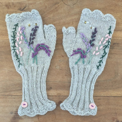 Paire de mitaines grises tricotées main, pour femme. Une composition florale évoquant de la bruyère est brodé en relief et est accompagnée de deux abeilles juste sous le feston supérieur. Le bord côte du poignet est long et se termine par un feston et un petit bouton rose pâle.