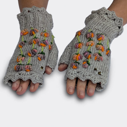 Mitaines grises tricotées à la main, présentées sur des mains à plat. Plusieurs tiges portent des fleurs multicolores en relief aux nuances de rose, de pêche et de mauve, relevées de touches orange vif.