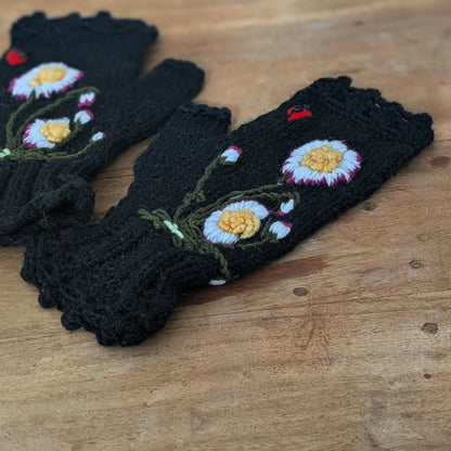 Photo d'une paire de mitaines femme artisanales posées à plat. La couleur est noire, en maille jersey, 2 pâquerettes sont brodées sur le dessus ainsi qu'une petite coccinelle rouge.