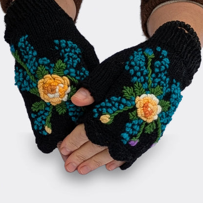 Mitaines noires tricotées main portées sur des mains légèrement croisées. Une fleur jaune nuancée de blanc et de doré est entourée de petites touches bleues disposées en étoile.