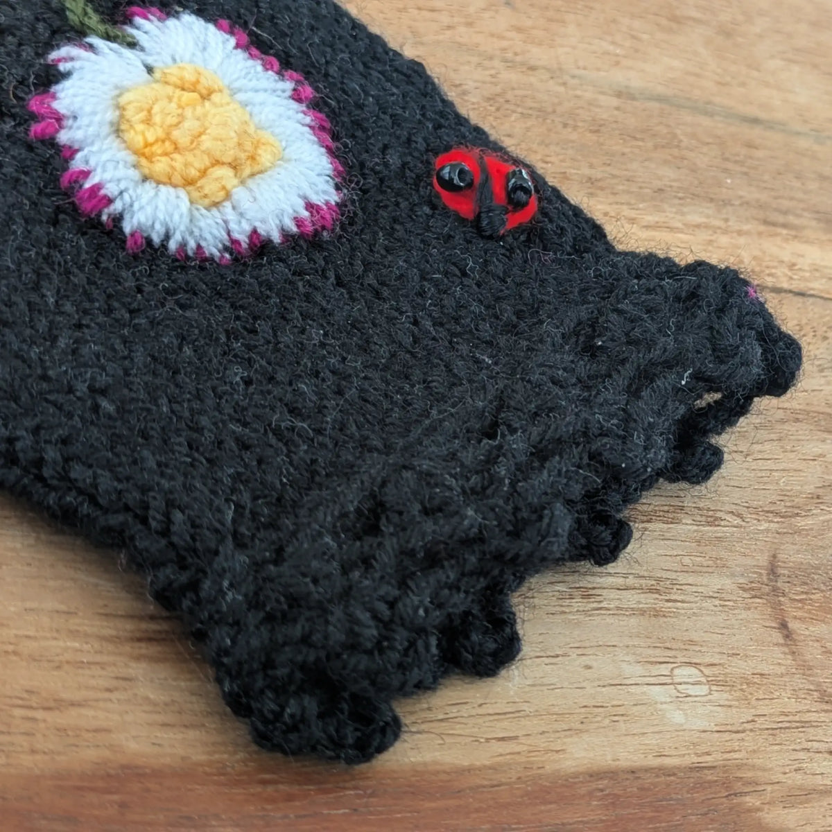 Vue en détail d'une paire de mitaines noire, la finition supérieure festonnée se détache nettement sur la photo ainsi que la composition florale et une petite coccinelle brodées sur le dessus.