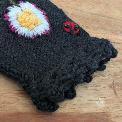 Vue en détail d'une paire de mitaines noire, la finition supérieure festonnée se détache nettement sur la photo ainsi que la composition florale et une petite coccinelle brodées sur le dessus.