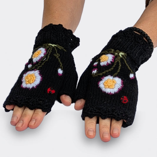 Mitaines pour femme noires présentées sur des mains à plat, avec deux pâquerettes en relief sur chaque mitaine. Les fleurs blanches aux cœurs jaunes sont entourées de boutons floraux et de tiges vert olive.