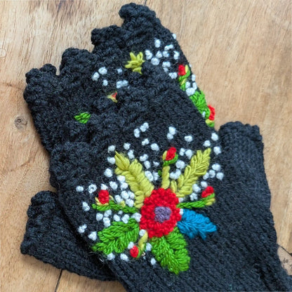 Mitaines noires tricotées main posées à plat, avec fleurs multicolores crochetées et broderies de feuillage.