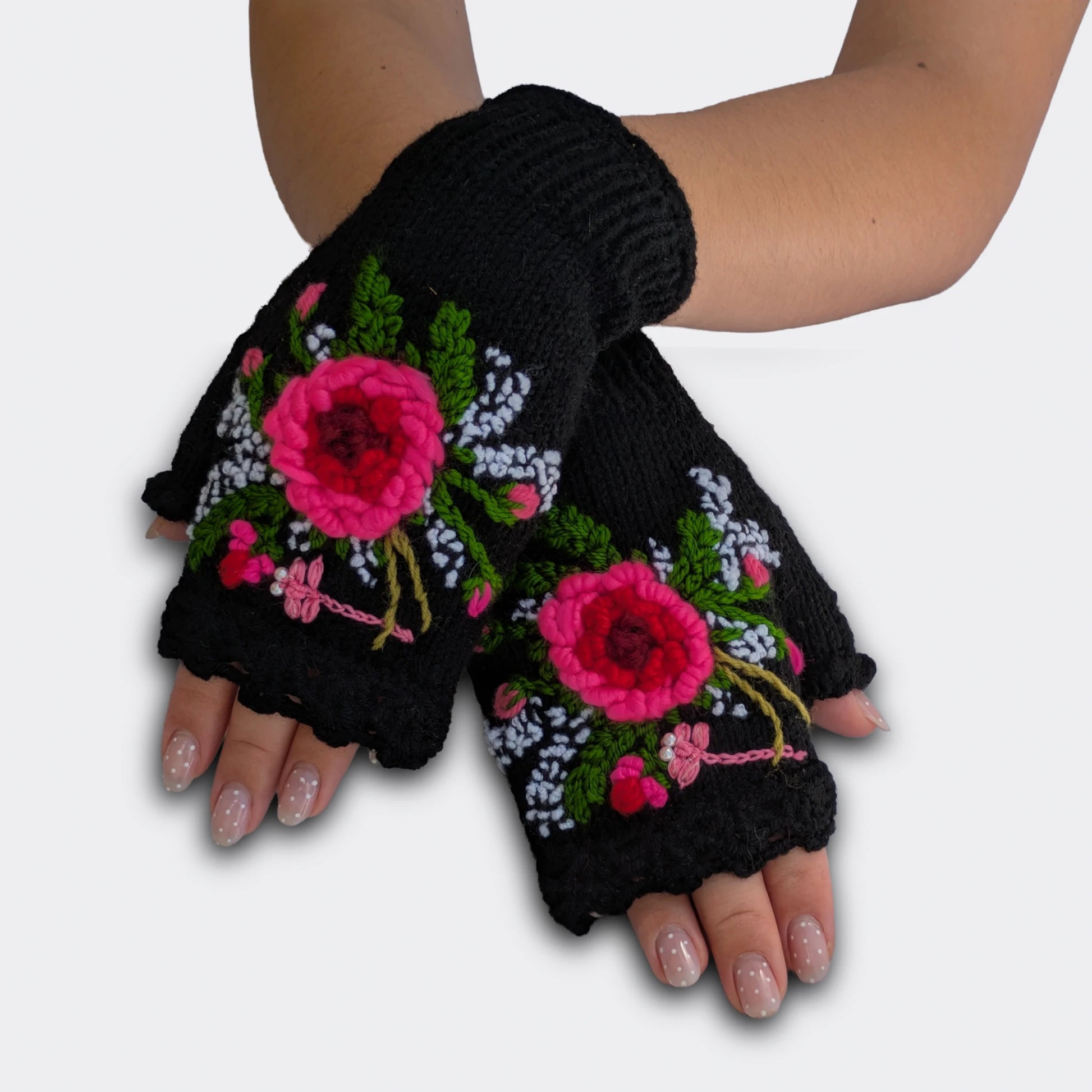 Mitaines faites main en Serbie, portées mains croisées. Une fleur rose en relief ressort sur la maille noire, accompagnée d’une libellule rose aux yeux perlés. Le haut des mitaines présente une finition festonnée.
