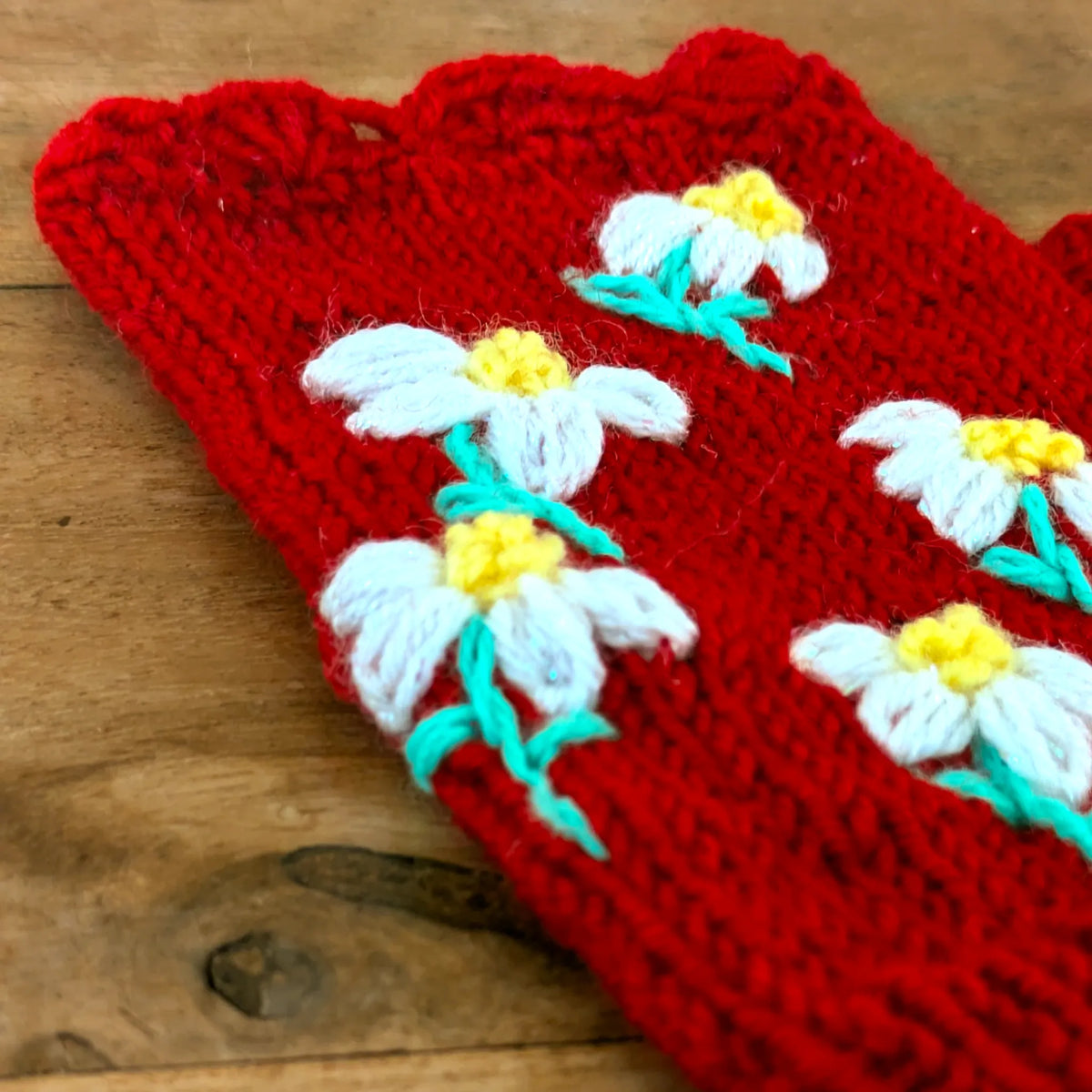 Vue d'une paire de mitaines rouges tricotées à la main. Elles sont dotées de cinq fleurettes jaunes et blanches. On y voit distinctement le bord supérieur festonnée.