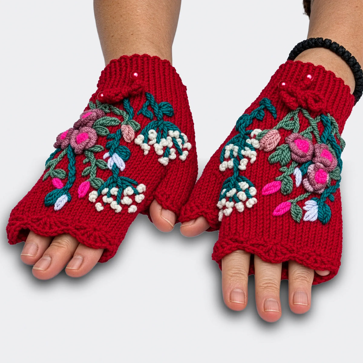 Mitaines rouges tricotées main présentées sur des mains à plat. Les roses en relief, les petites fleurs blanches et le feuillage vert bleuté se déploient sur la maille jersey.
