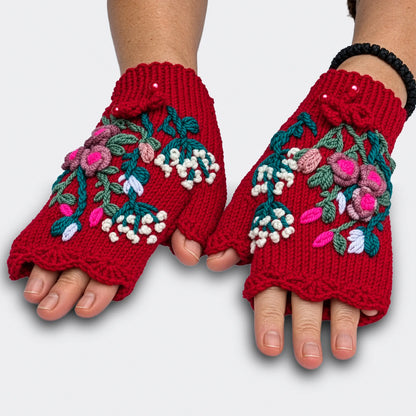 Mitaines rouges tricotées main présentées sur des mains à plat. Les roses en relief, les petites fleurs blanches et le feuillage vert bleuté se déploient sur la maille jersey.