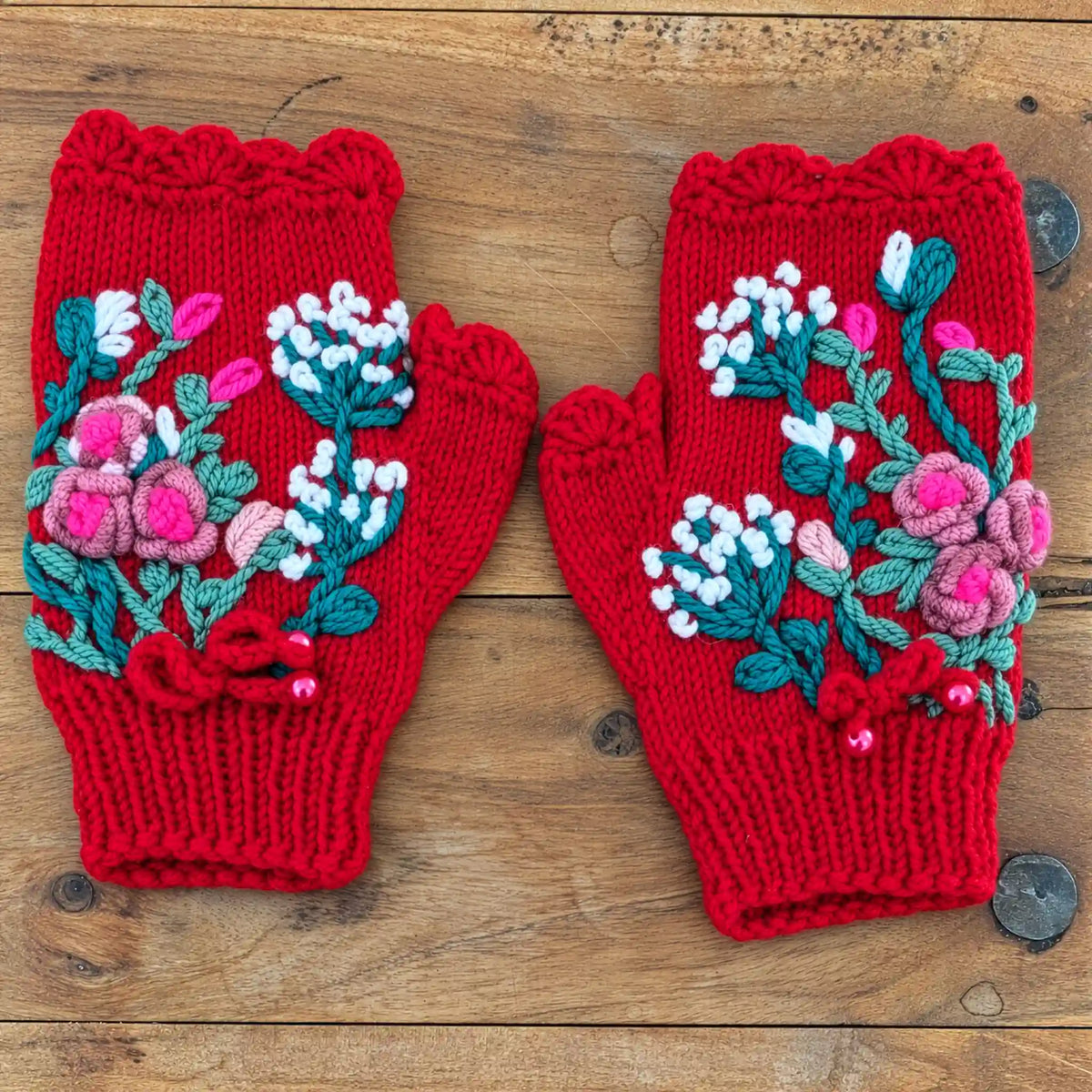 Mitaines rouges avec des une composition florale crochetée puis brodée sur la mitaine. Petites roses bouton et fleurs blanches. Vue à plat sur une table.