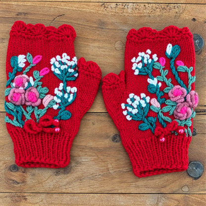 Mitaines rouges avec des une composition florale crochetée puis brodée sur la mitaine. Petites roses bouton et fleurs blanches. Vue à plat sur une table.