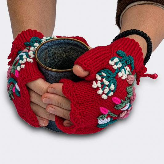 Des mains tenant une tasse portent des mitaines artisanales rouges ornées d'un bouquet floral. Les fleurs roses, les touches de blanc et les feuilles brodées composent un décor en relief.