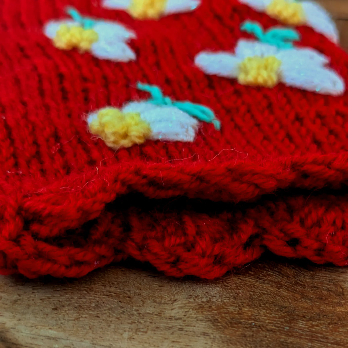 Détail du bord supérieur festonné d'une paire de mitaines rouges avec des petites fleurs blanches et jaunes.