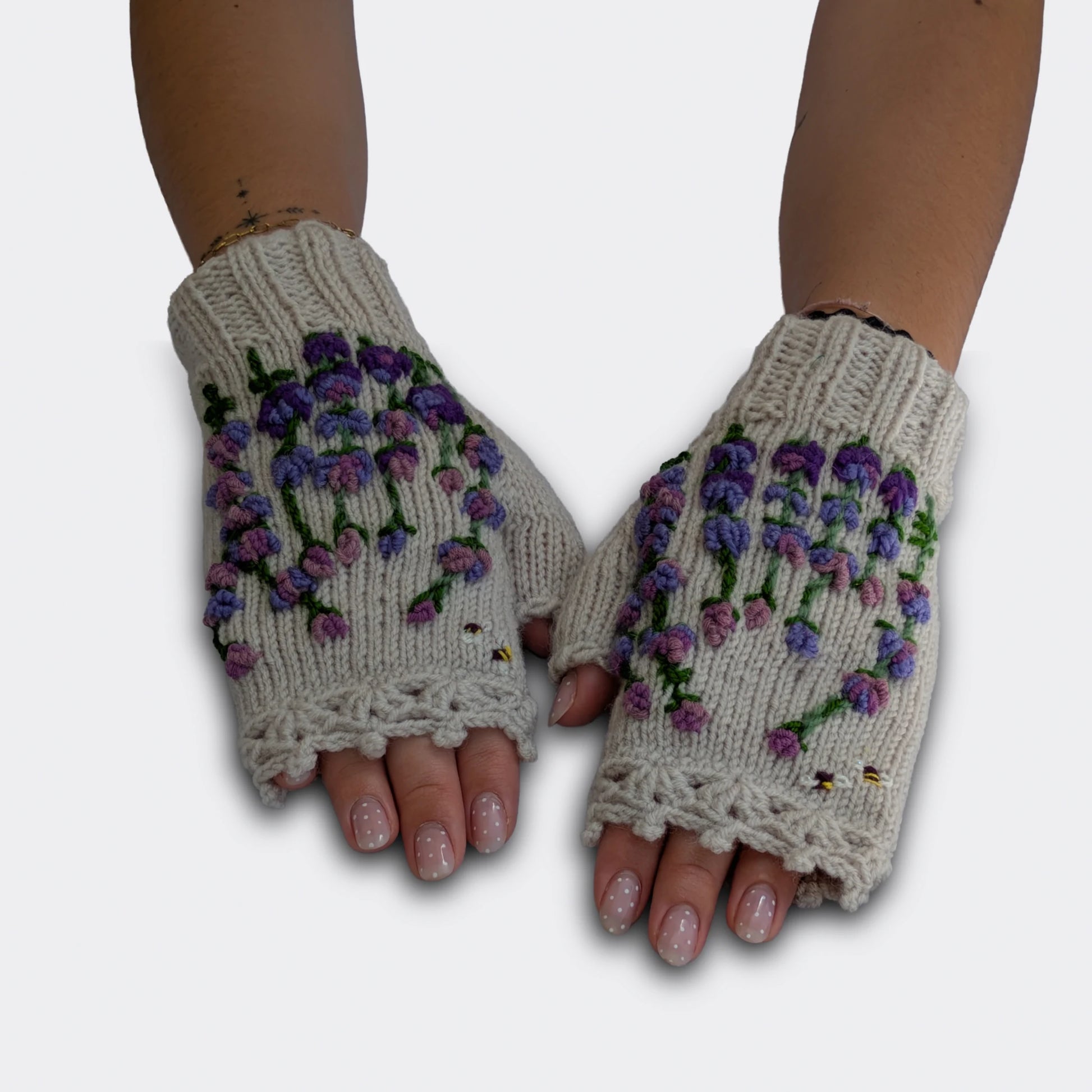 Mitaines gris clair tricotées main, portées à plat. Le décor montre de petites fleurs mauves, violettes et parme en grappes, avec tiges vertes. Deux petites abeilles complètent la composition.