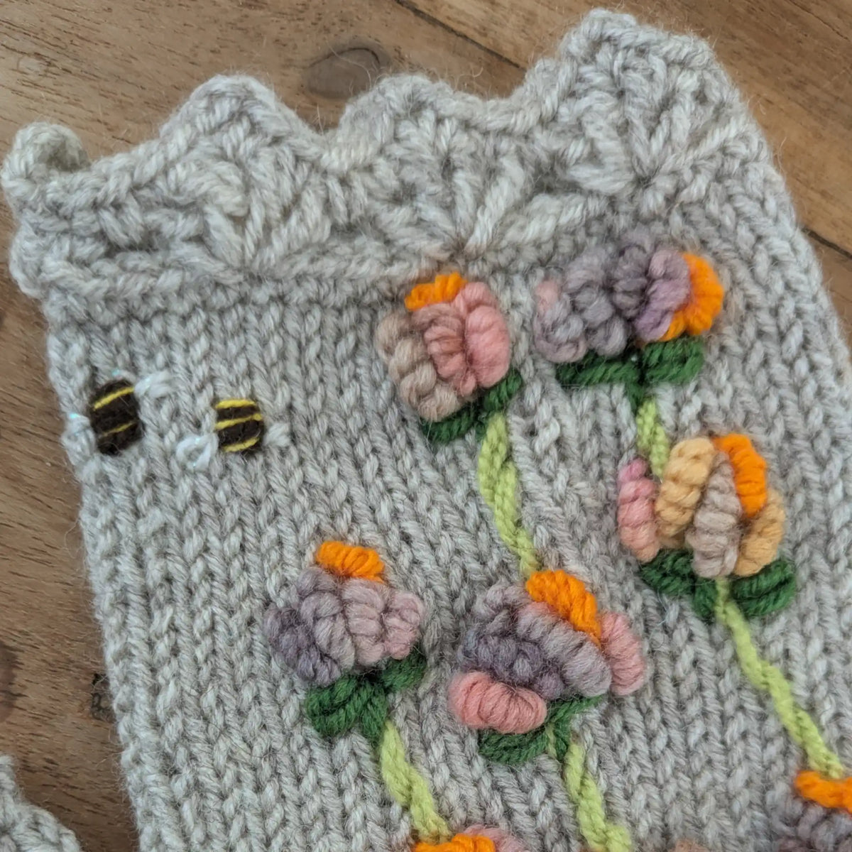 Vue rapprochée du bord supérieur festonné d'une mitaine artisanale grise. De petites abeilles brodées voisinent avec des fleurs stylisées roses, mauves et orangées en relief.