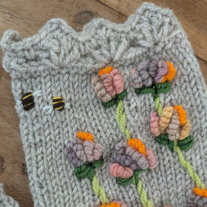 Vue rapprochée du bord supérieur festonné d'une mitaine artisanale grise. De petites abeilles brodées voisinent avec des fleurs stylisées roses, mauves et orangées en relief.