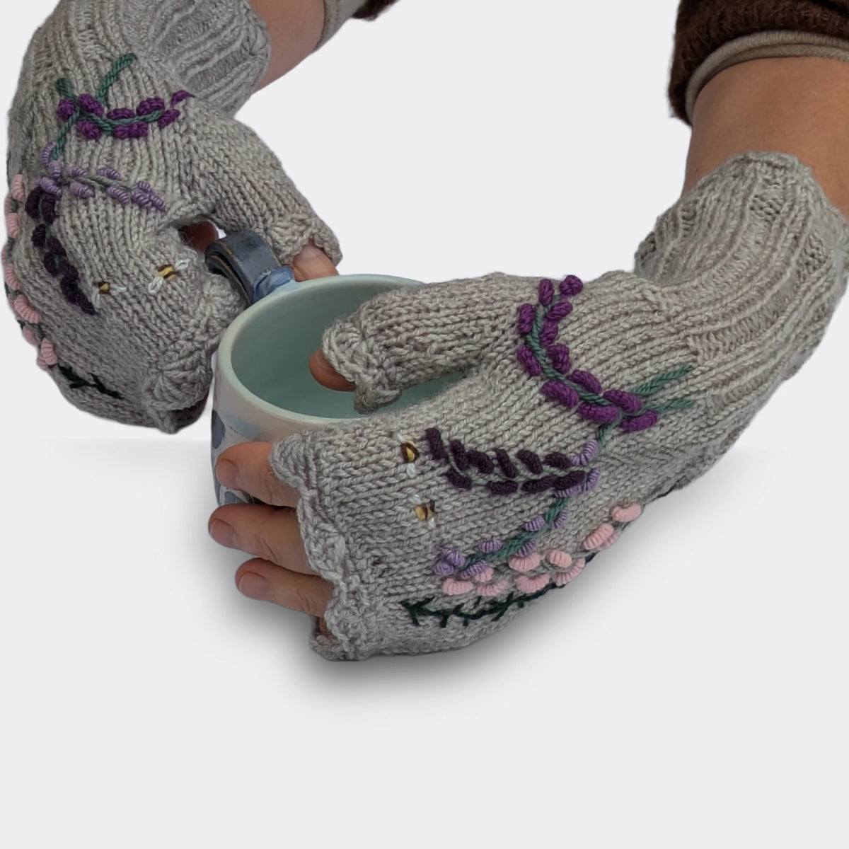 Deux mains légèrement croisées présentent des mitaines tricotées main, ornées de fleurs aux tons roses et mauves. Les poignets particulièrement longs, travaillés en côtes, prolongent la maille jersey grise.