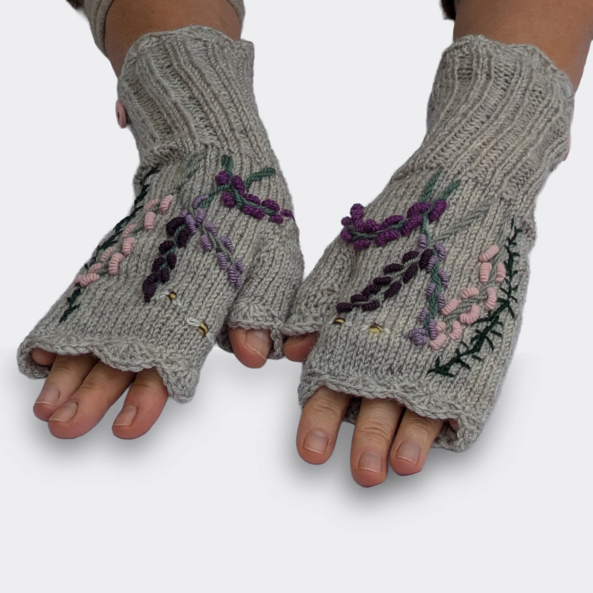 Paire de mitaines artisanales grises à composition florale, portée sur des mains à plat. Les fines tiges fleuries se détachent au-dessus des longs poignets en côtes, avec des bordures festonnées.