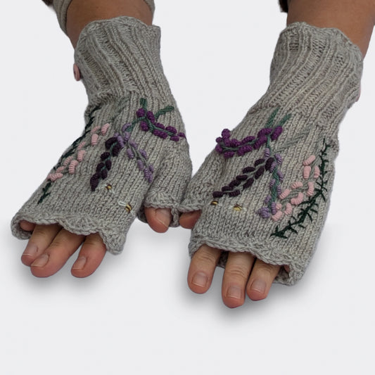 Paire de mitaines artisanales grises à composition florale, portée sur des mains à plat. Les fines tiges fleuries se détachent au-dessus des longs poignets en côtes, avec des bordures festonnées.