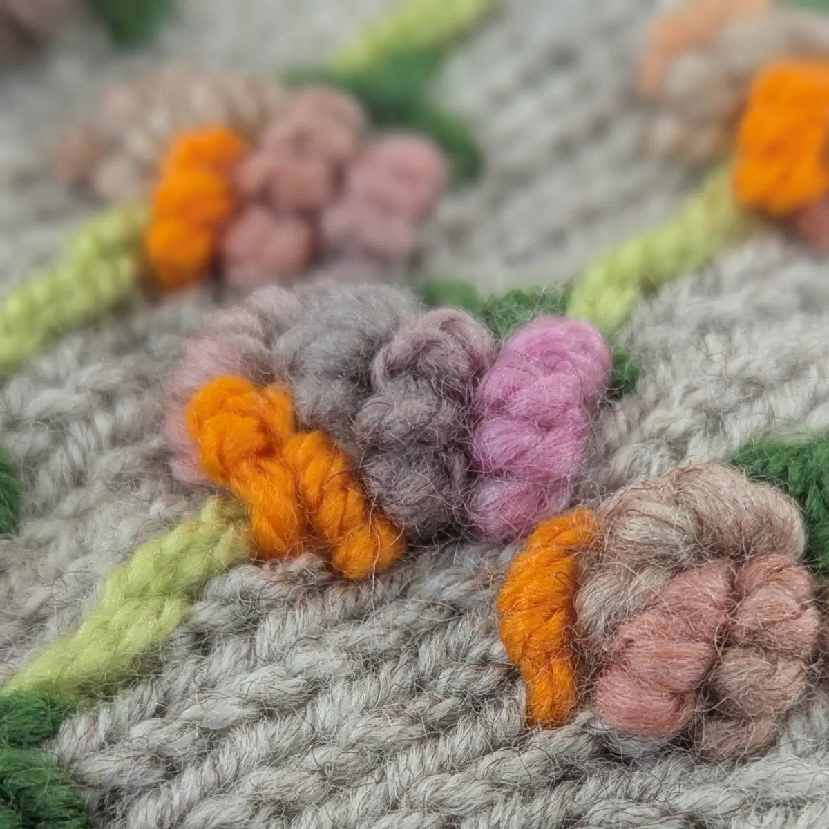 Gros plan sur les fleurs en relief de mitaines pour femme grises. Les fils rose poudré, lilas, pêche et orange vif forment de petites corolles arrondies, disposées sur des tiges vert clair.