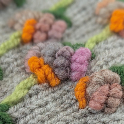 Gros plan sur les fleurs en relief de mitaines pour femme grises. Les fils rose poudré, lilas, pêche et orange vif forment de petites corolles arrondies, disposées sur des tiges vert clair.