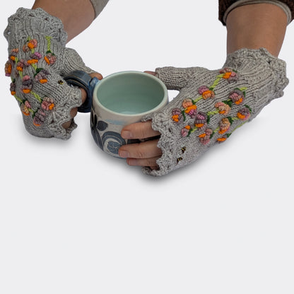 Deux mains tenant une tasse portent des mitaines grises à fleurs multicolores en relief. De petites abeilles et des tiges garnies de fleurs aux touches orange vif animent le tricot.