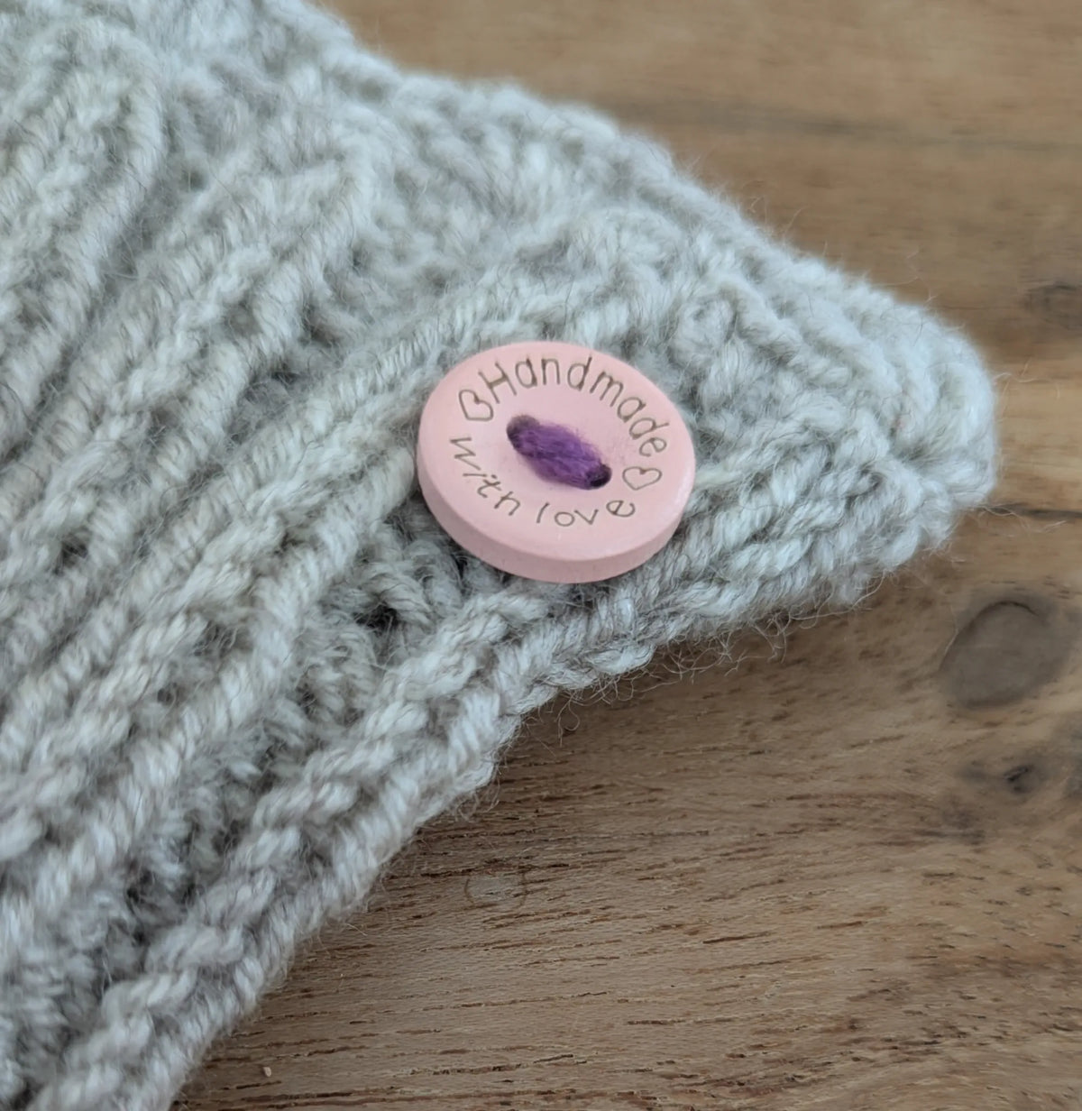 Détail du bord côte d'une paire de mitaine grise. On y voit un petit bouton rose pâle ajoutant une touche de fantaisie.