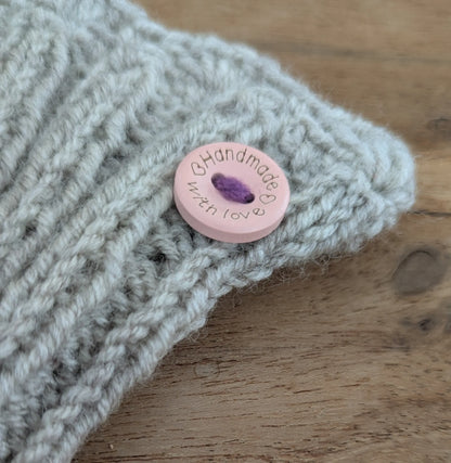 Détail du bord côte d'une paire de mitaine grise. On y voit un petit bouton rose pâle ajoutant une touche de fantaisie.