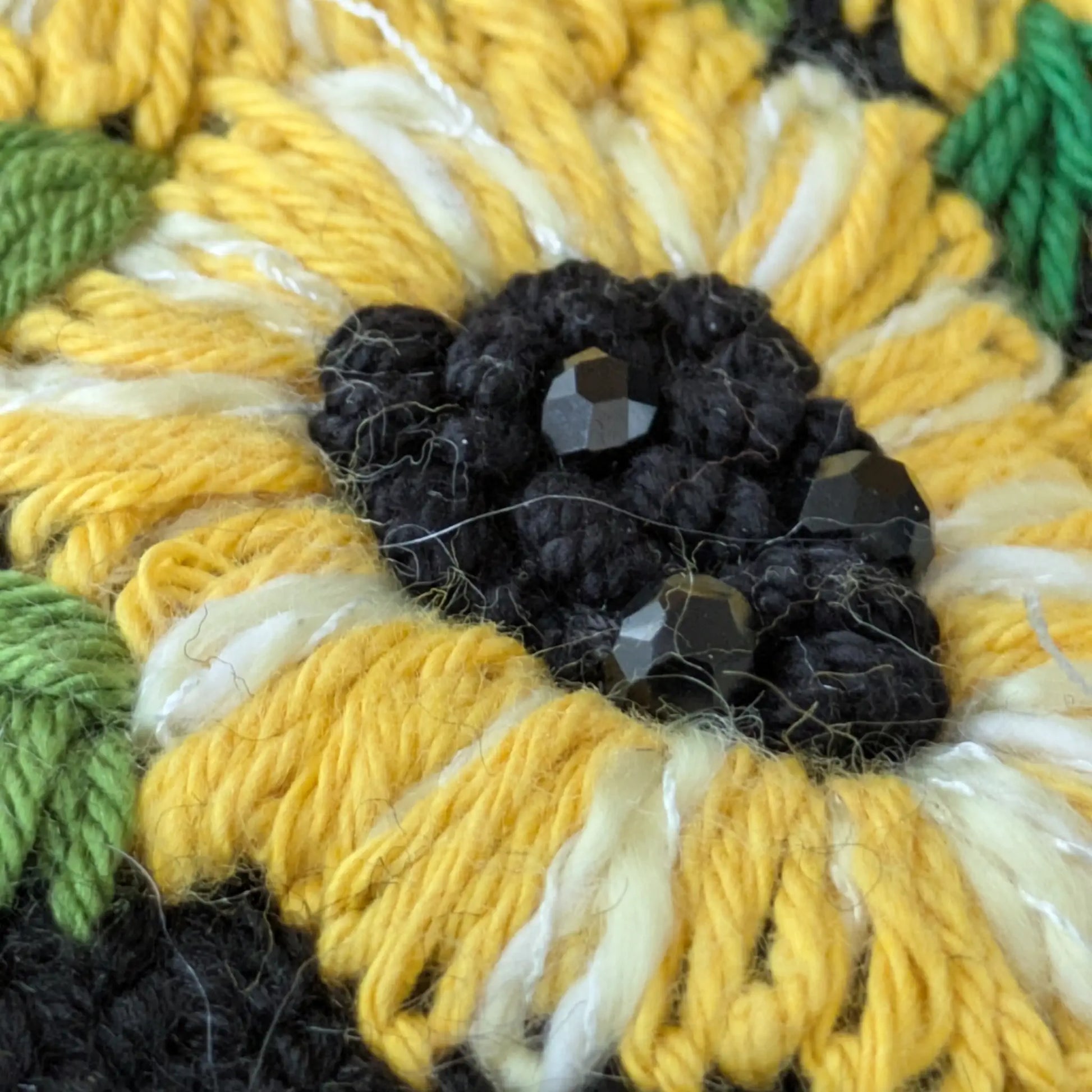 Détail d’un tournesol jaune en relief avec cœur en perles noires sur des mitaines noires.