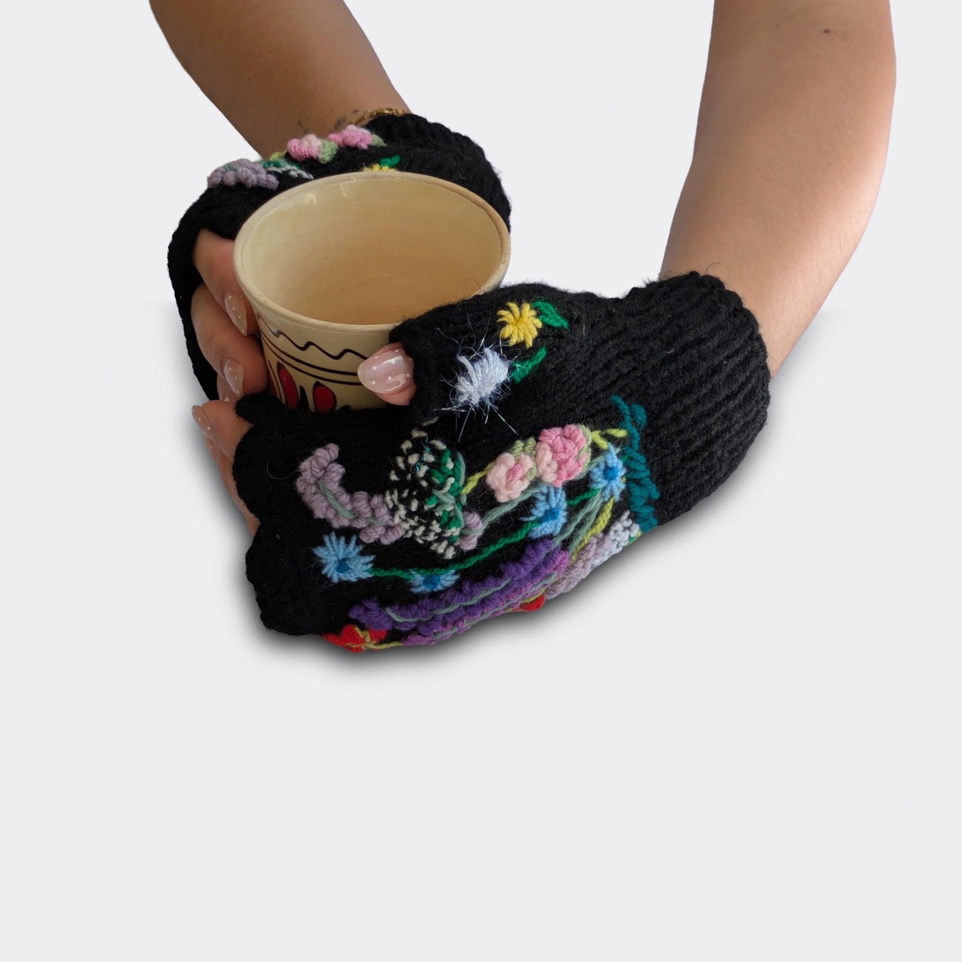 Mitaines noires tenant une tasse, décorées de fleurs multicolores brodées et d’un bord supérieur festonné.