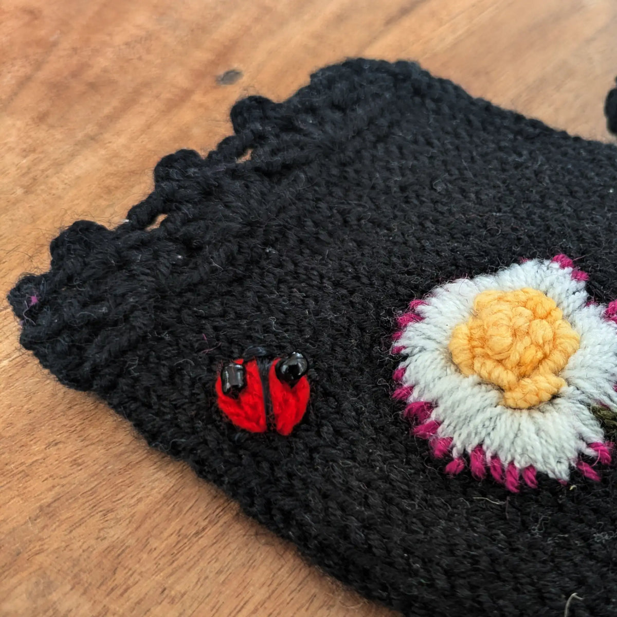 Vue en détail du feston d'une paire artisanale de mitaine noire en jersey ainsi que du décor floral et d'une petite coccinelle brodée sur le dessus.