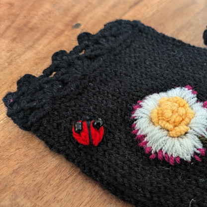 Vue en détail du feston d'une paire artisanale de mitaine noire en jersey ainsi que du décor floral et d'une petite coccinelle brodée sur le dessus.
