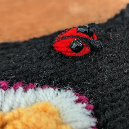 Vue en très gros plan d'une coccinelle rouge brodée sur le dessus d'une mitaine noire en jersey. Les boutons sur le dos sont mis en valeur avec deux perles noires brillantes.