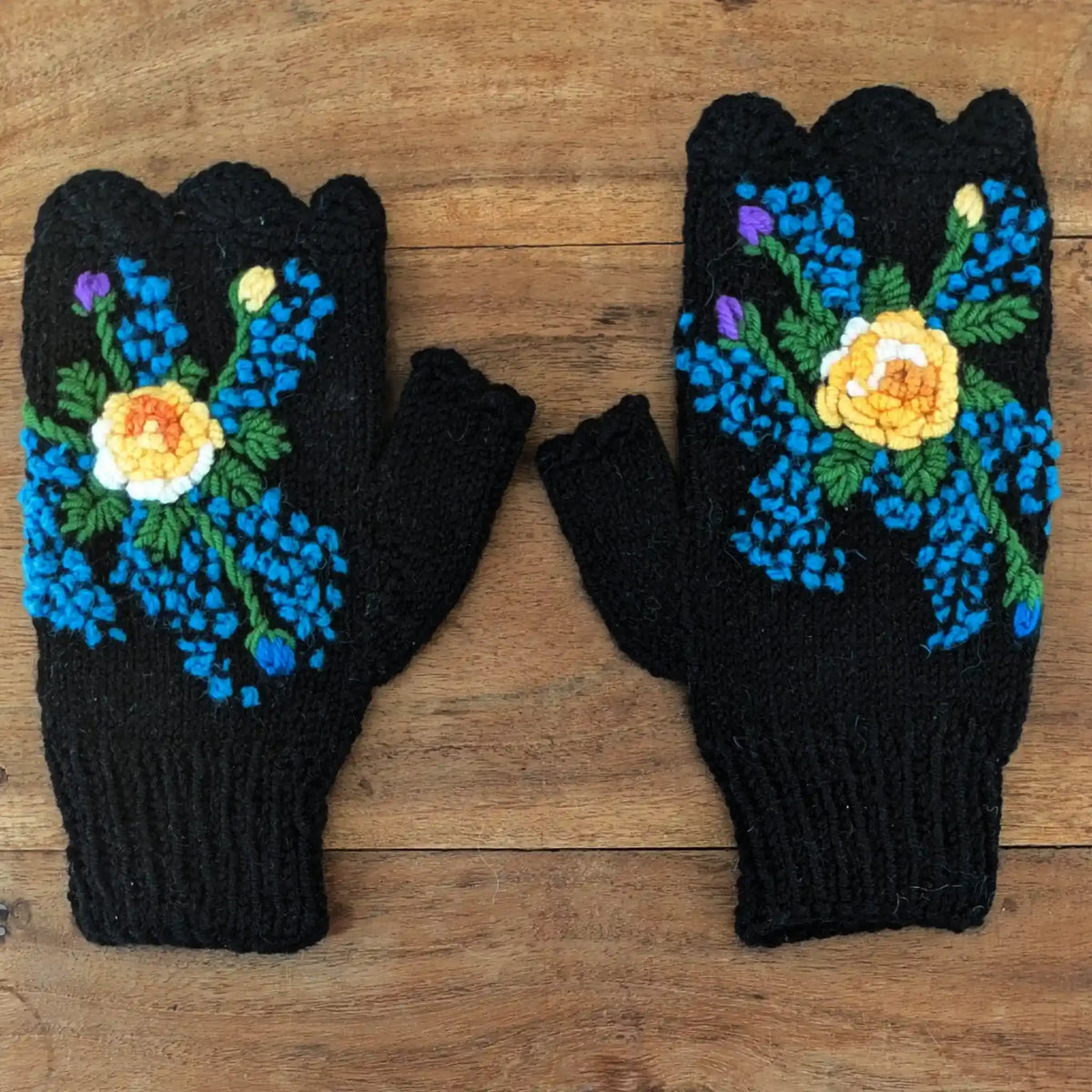 Mitaines pour femmes posées à plat sur une table en bois, elles sont noires avec une fleur centrale jaune crochetée en relief et de petits points bleus tout autour.