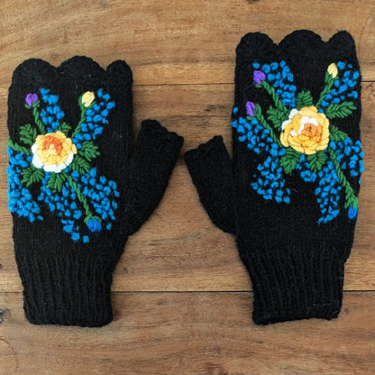 Mitaines pour femmes posées à plat sur une table en bois, elles sont noires avec une fleur centrale jaune crochetée en relief et de petits points bleus tout autour.