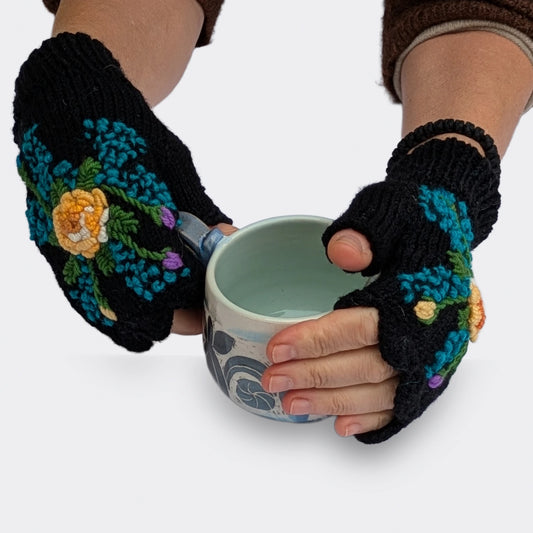 Deux mains tenant une tasse portent des mitaines noires tricotées à la main. Les fleurs centrales jaune doré et les petites touches bleues ressortent sur la maille noire.