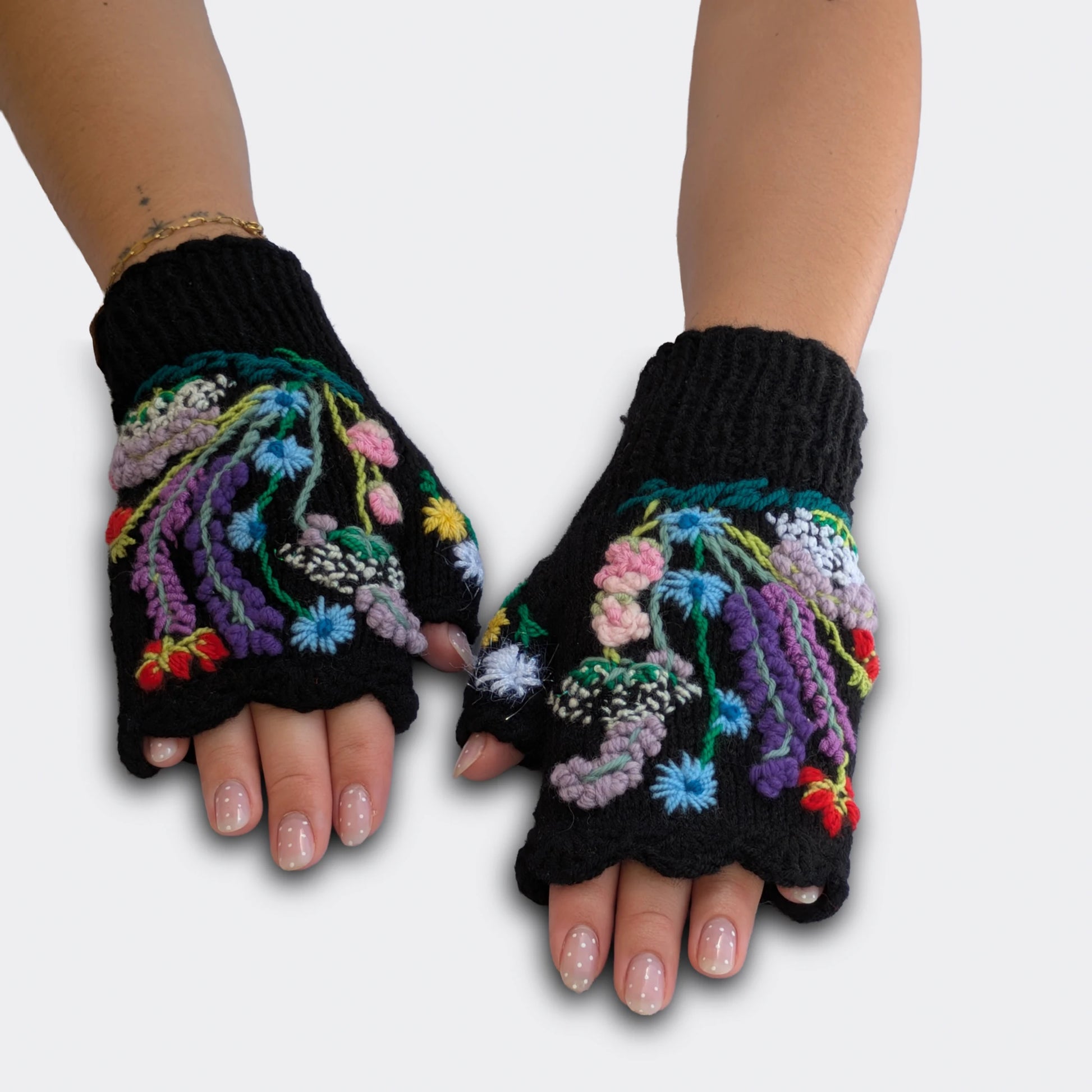 Mitaines noires tricotées main, portées à plat, avec fleurs brodées multicolores sur le dessus des mains.